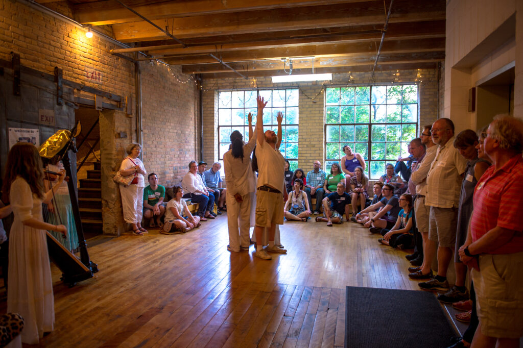 A group of people stands together in the middle of a circle of onlookers, beautifully backlit, reaching toward the heavens.