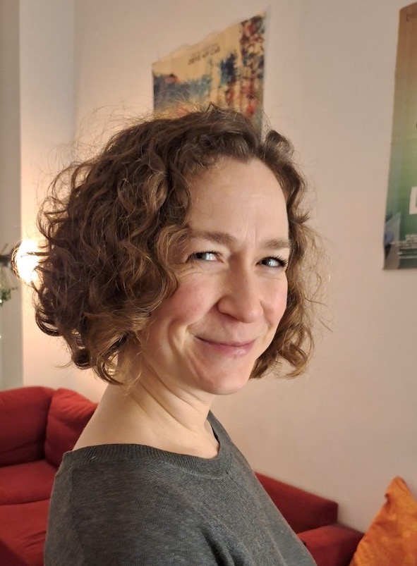 A white woman with short curly hair wearing a grey sweater smiles over her shoulder at the camera.